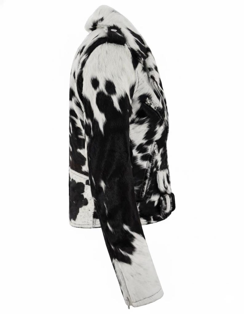 May include: A side view of a moto-style jacket featuring a black and white cow-print pattern. The jacket includes a notched collar, long sleeves with zippered cuffs, a silver-toned front zipper, and a buckled belt detail at the waist hem.