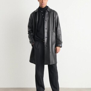 May include: A full-length image of a person wearing a black leather trench coat over a black polo shirt and black trousers. The coat has a button-up front and a collared neckline. The person is also wearing black shoes.