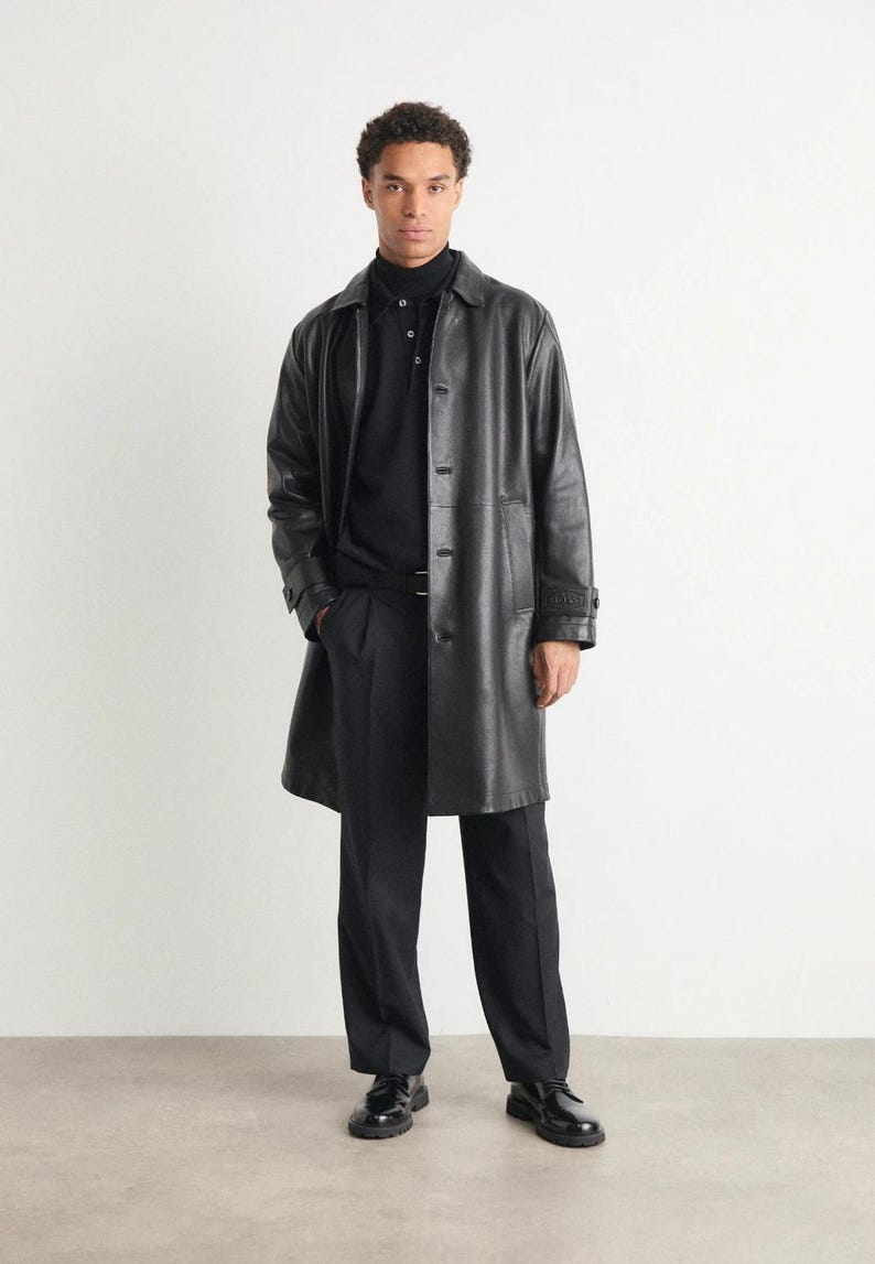 May include: A full-length image of a person wearing a black leather trench coat over a black polo shirt and black trousers. The coat has a button-up front and a collared neckline. The person is also wearing black shoes.