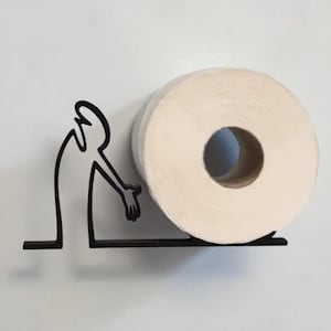 May include: A black metal toilet paper holder featuring a stylized figure reaching towards a roll of white toilet paper. The holder is mounted on a white wall, creating a minimalist and modern bathroom accessory.