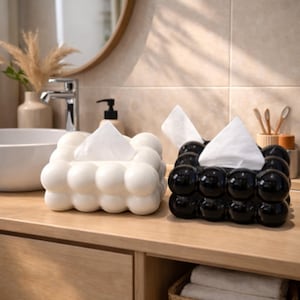 May include: Two decorative tissue box holders, one white and one black, sit side-by-side on a wooden surface. Each holder has a bubble-like design and dispenses tissues from the top. The bathroom setting includes a sink, mirror, and other accessories.