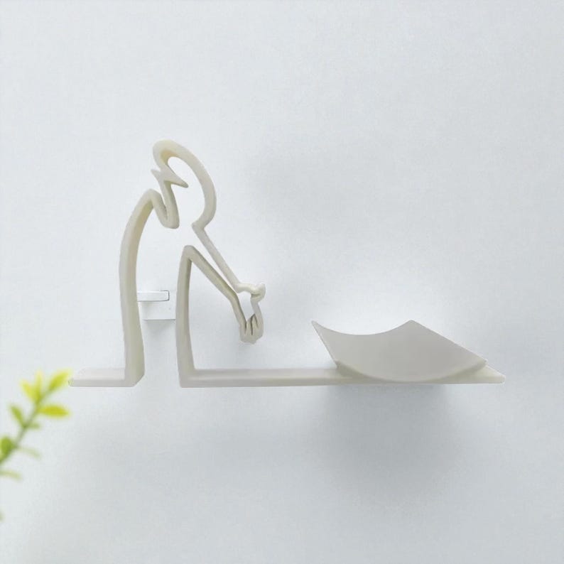 May include: A minimalist, off-white wall shelf featuring a stylized human figure leaning over a small, curved tray. The shelf is mounted on a white wall, with a simple, modern design.