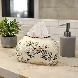 Puede incluir: Caja de pañuelos de cerámica color crema con un estampado floral en rosa y verde. La caja de pañuelos tiene un borde festoneado con un ribete rojo oscuro. También se ve un dispensador de jabón gris y una pequeña planta en maceta.