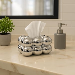Peut inclure: Un distributeur de mouchoirs chromé avec un design de bulles, contenant des mouchoirs blancs. Un distributeur de savon gris avec une pompe argentée est à proximité. Une petite plante en pot est également visible.