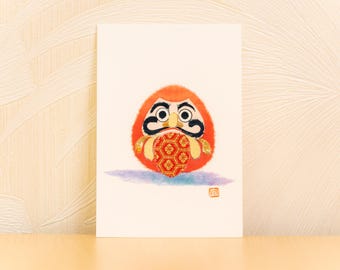 Könnte beinhalten: Illustration einer Daruma-Puppe auf einer weißen Karte. Die Puppe ist orange mit schwarzen Gesichtszügen und einem rot-gold gemusterten Objekt. Ein kleiner roter Stempel befindet sich unten rechts. Der Hintergrund ist hellbeige.