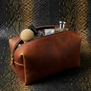 May include: A brown leather wash bag with a partially open zip, displaying grooming products. The rectangular bag has rounded corners and a brass zip. A makeup brush, perfume bottle, and other items are visible inside. The background has a snakeskin pattern.