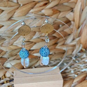 Puede incluir: Pendientes colgantes con tachuelas circulares doradas. Cada pendiente presenta una cuenta estampada en azul y blanco, una cuenta de cristal transparente y una cuenta rectangular blanca, conectadas con alambre dorado.