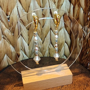 Puede incluir: Un par de pendientes colgantes con ganchos dorados y una serie de cuentas transparentes y doradas. Los pendientes se exhiben en un soporte de acrílico transparente con una base de madera, sobre un fondo tejido.