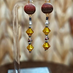 May include: A pair of dangle earrings with a red and brown circular top, black beads, white beads, silver beads, and two yellow diamond-shaped beads. The earrings are displayed on a clear stand.