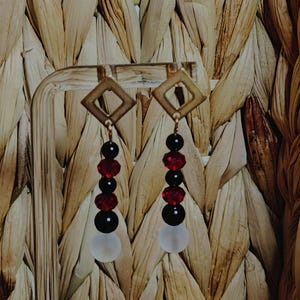 May include: A pair of dangle earrings with a geometric square top. The earrings feature a combination of black, red, and white beads. The earrings are suspended from a gold-colored metal hook.