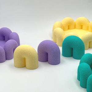 Op de afbeelding: Een verzameling kleurrijke, getextureerde, U-vormige objecten in tinten paars, geel en blauwgroen. Sommige zijn in een cirkelvorm gerangschikt, andere staan ​​alleen. De objecten hebben een zachte, afgeronde uitstraling.