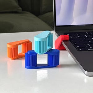 Peut inclure: Plusieurs supports d'ordinateur portable colorés en orange, bleu, bleu clair et rouge. Les supports sont fabriqués dans un matériau nervuré et sont conçus pour soutenir un ordinateur portable. Un support rouge est fixé à un ordinateur portable.