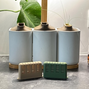 Könnte beinhalten: Drei hellblaue Keramikvasen mit braunen Oberteilen stehen auf einem Holzsockel. Zwei kleine, rechteckige Lufterfrischer, einer beige und einer grün, sind davor. Eine klare Glasflasche mit einem Pumpspender befindet sich rechts.