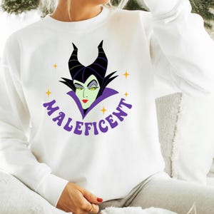 Puede incluir: Sudadera blanca con un dise&ntilde;o de Mal&eacute;fica, la villana de Disney. El dise&ntilde;o incluye la cara de Mal&eacute;fica con cuernos negros, un cuello morado y la palabra "Mal&eacute;fica" en morado. Acentos de estrellas amarillas rodean la imagen.