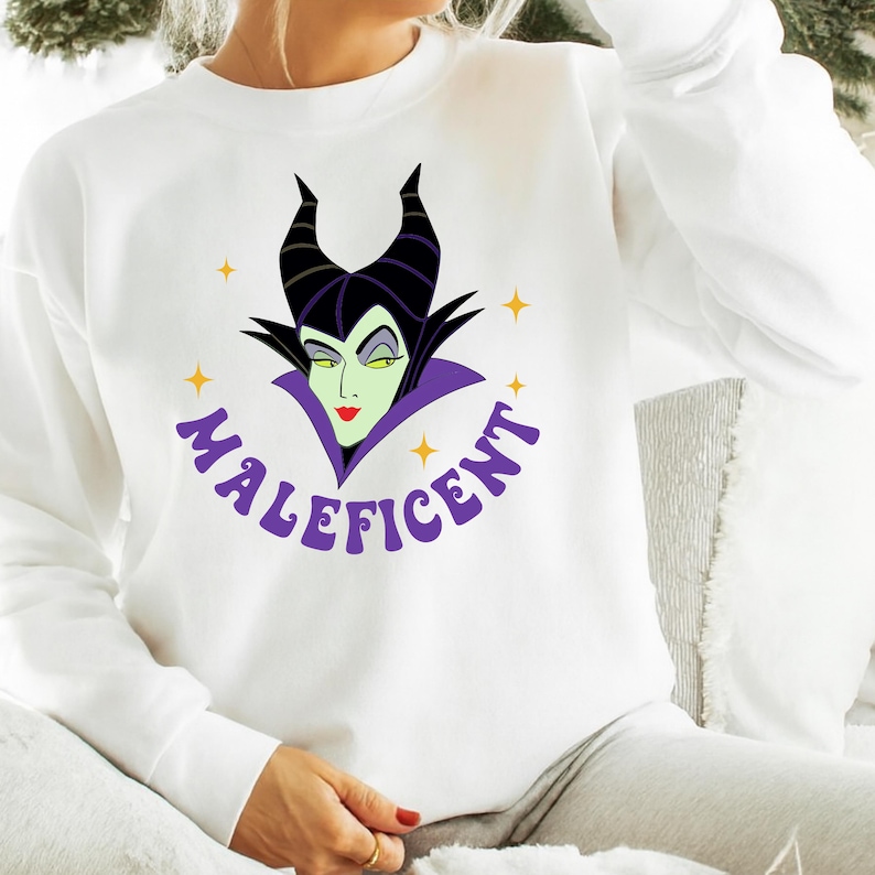 Puede incluir: Sudadera blanca con un dise&ntilde;o de Mal&eacute;fica, la villana de Disney. El dise&ntilde;o incluye la cara de Mal&eacute;fica con cuernos negros, un cuello morado y la palabra "Mal&eacute;fica" en morado. Acentos de estrellas amarillas rodean la imagen.