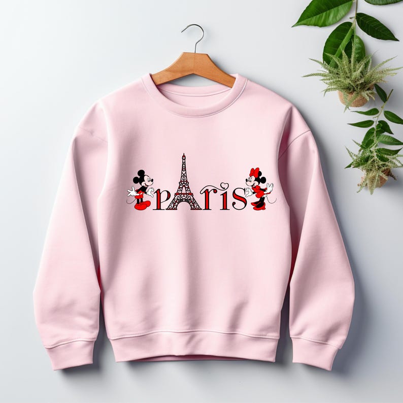 May include: A pale pink sweatshirt with the word "Paris" and the Eiffel Tower in the centre. Mickey and Minnie Mouse are on either side of the word. The sweatshirt is displayed on a wooden hanger.