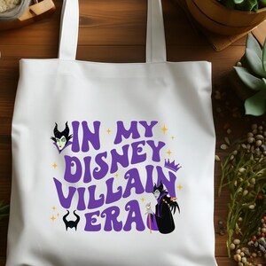 Puede incluir: Bolso tote blanco con el texto "IN MY DISNEY VILLAIN ERA" en morado, e ilustraciones de villanos de Disney. El bolso tiene dos asas y est&aacute; hecho de una tela resistente.