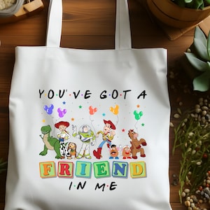 May include: White tote bag featuring a colorful graphic of characters and the text "YOU'VE GOT A FRIEND IN ME." The design includes cartoon figures, balloons, and stars. The bag has two white handles.