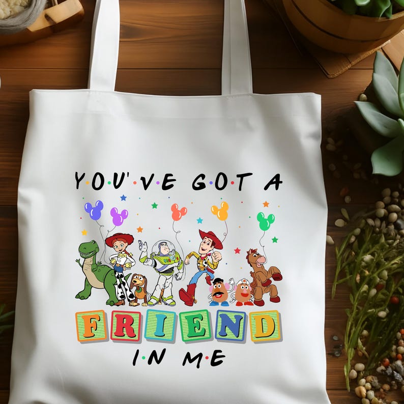 May include: White tote bag featuring a colorful graphic of characters and the text "YOU'VE GOT A FRIEND IN ME." The design includes cartoon figures, balloons, and stars. The bag has two white handles.