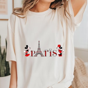 May include: A cream-coloured t-shirt with a graphic of Mickey and Minnie Mouse, the Eiffel Tower, and the word "Paris" in red and black. Mickey and Minnie are in their classic outfits. The shirt is a casual style.