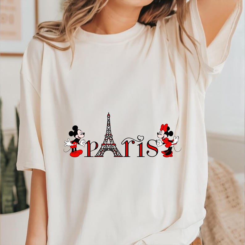 May include: A cream-coloured t-shirt with a graphic of Mickey and Minnie Mouse, the Eiffel Tower, and the word "Paris" in red and black. Mickey and Minnie are in their classic outfits. The shirt is a casual style.