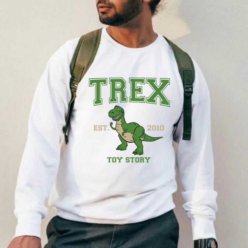 K&ouml;nnte beinhalten: Wei&szlig;es Langarm-Sweatshirt mit einem gr&uuml;nen Cartoon-T-Rex-Dinosaurier-Motiv. Das Wort "TREX" ist &uuml;ber dem Dinosaurier aufgedruckt, mit "EST. 2010" und "TOY STORY" darunter. Das Sweatshirt hat einen Rundhalsausschnitt.