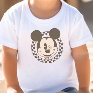 Op de afbeelding: Wit T-shirt met een lachende Mickey Mouse-afbeelding. De afbeelding toont Mickey's gezicht, oren en een geblokte rand. Het T-shirt heeft korte mouwen en lijkt van zacht materiaal te zijn.