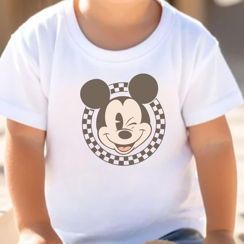Op de afbeelding: Wit T-shirt met een lachende Mickey Mouse-afbeelding. De afbeelding toont Mickey's gezicht, oren en een geblokte rand. Het T-shirt heeft korte mouwen en lijkt van zacht materiaal te zijn.