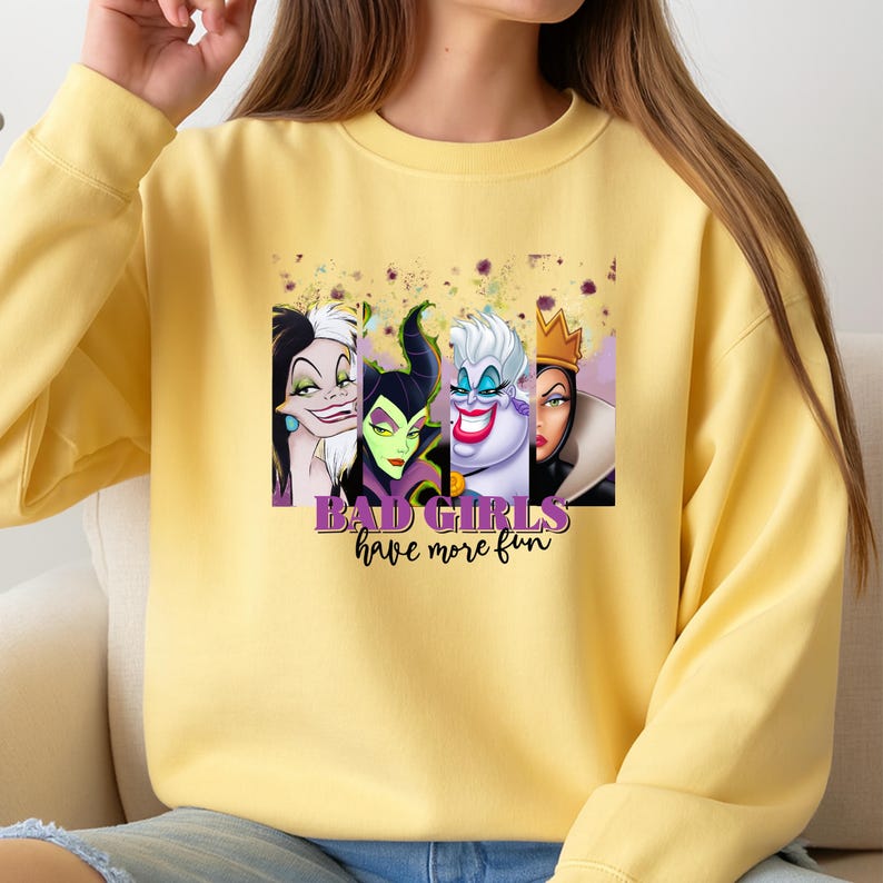 Puede incluir: Una sudadera de cuello redondo en color amarillo p&aacute;lido con un estampado gr&aacute;fico de cuatro villanas animadas dispuestas en paneles verticales. Debajo de los personajes, el texto dice "BAD GIRLS have more fun" en letras moradas y negras.