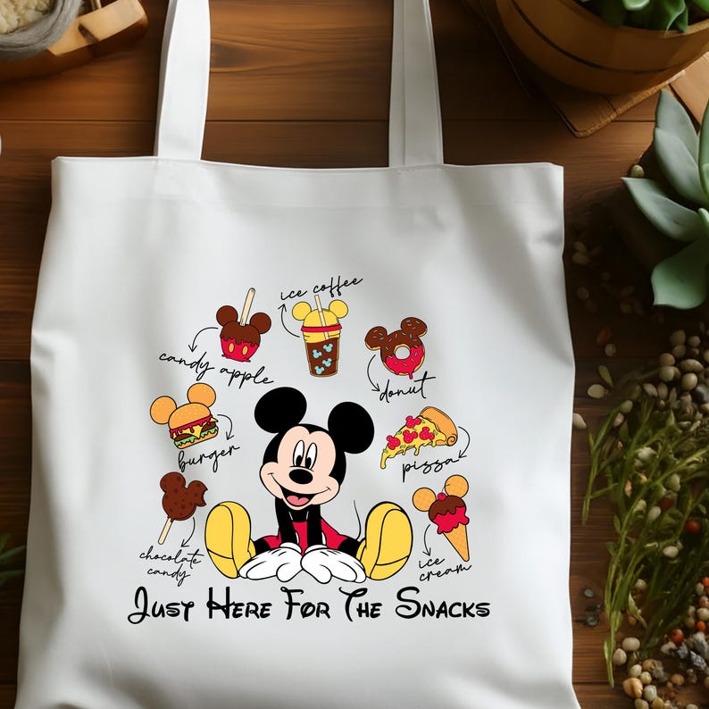 Puede incluir: Bolso tote blanco con Mickey Mouse y varias ilustraciones de comida, incluyendo una manzana de caramelo, hamburguesa, caf&eacute; helado, donut, pizza, bomb&oacute;n de chocolate y helado. El texto "JUST HERE FOR THE SNACKS" est&aacute; impreso debajo.