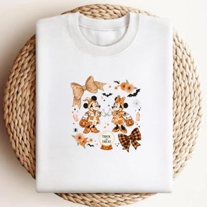 Op de afbeelding: Witte sweatshirt met een Halloween-thema met Mickey en Minnie Mouse in kostuums, met pompoenen. Het ontwerp bevat oranje bloemen, vleermuizen en de woorden "Trick or Treat". Twee grote strikken maken het ontwerp compleet.