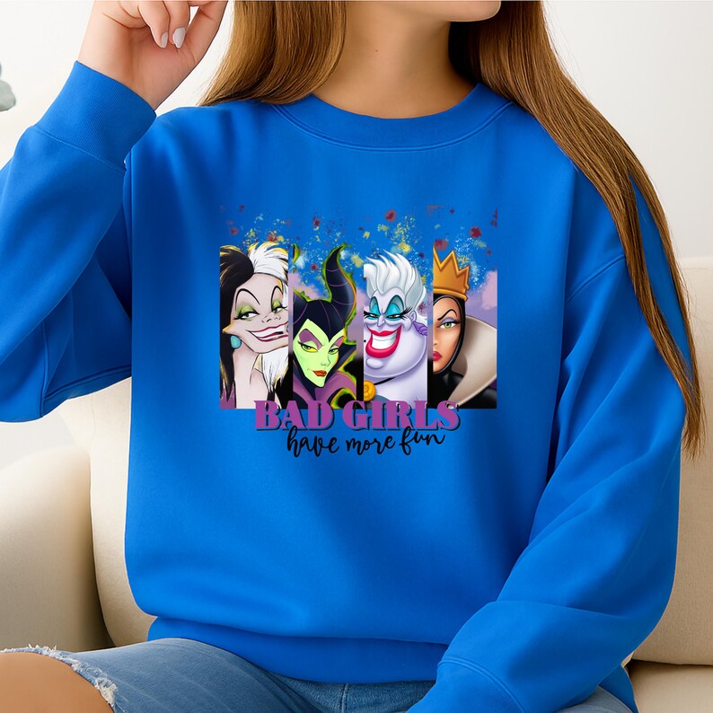 Puede incluir: Una sudadera azul brillante de cuello redondo con un estampado gr&aacute;fico de cuatro villanas animadas. El texto "BAD GIRLS have more fun" est&aacute; impreso en morado y negro debajo de los personajes. El dise&ntilde;o incluye a Cruella de Vil, Mal&eacute;fica, &Uacute;rsula y la Reina Malvada.