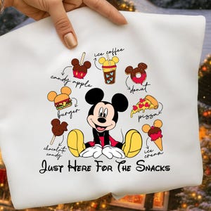 Puede incluir: Camiseta blanca con Mickey Mouse rodeado de dibujos de snacks como una manzana de caramelo, hamburguesa y pizza. El texto "Just Here For The Snacks" est&aacute; debajo del personaje. El dise&ntilde;o incluye ilustraciones coloridas de varias golosinas.