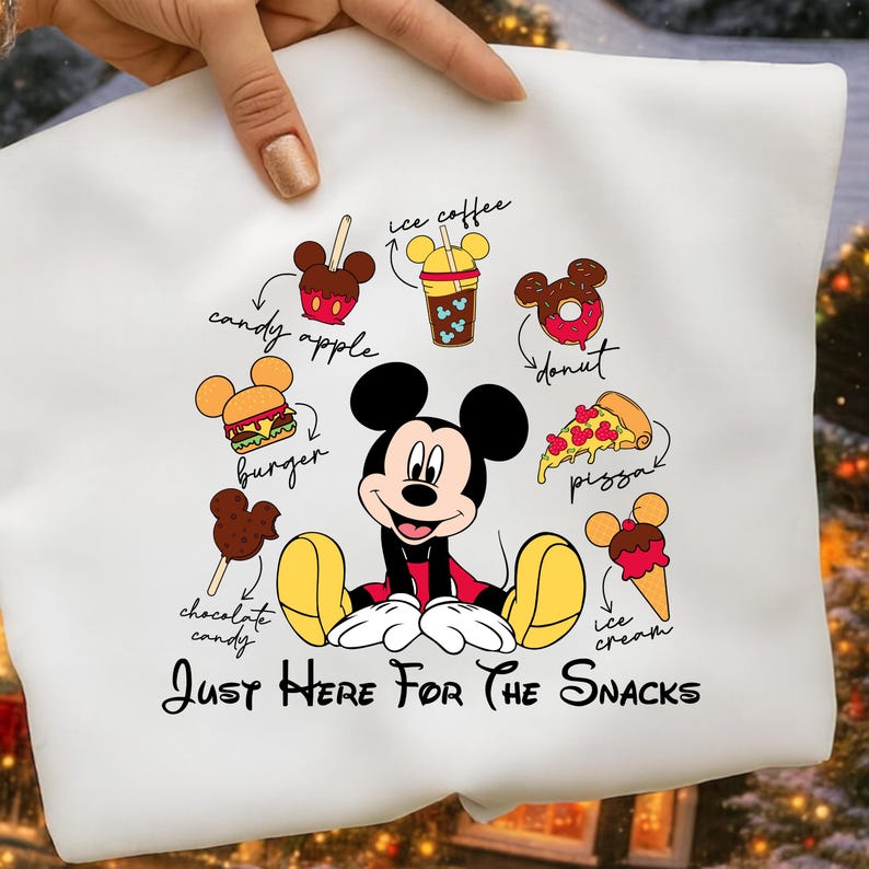 Puede incluir: Camiseta blanca con Mickey Mouse rodeado de dibujos de snacks como una manzana de caramelo, hamburguesa y pizza. El texto "Just Here For The Snacks" est&aacute; debajo del personaje. El dise&ntilde;o incluye ilustraciones coloridas de varias golosinas.
