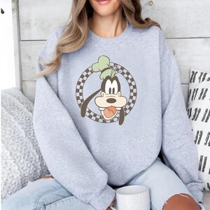 Op de afbeelding: Een lichtgrijs sweatshirt met een cartoonafbeelding van Goofy. De afbeelding toont Goofy's gezicht met een groene hoed, in een bruine en witte geruite cirkel. De sweatshirt is een crew neck stijl.