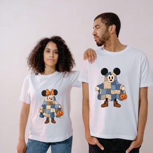 May include: White t-shirts with a cartoon character dressed as a ghost in a patchwork quilt. The character is holding a jack-o'-lantern. The character has large black ears and brown boots. The other character has a bow.
