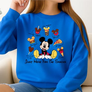 Puede incluir: Una sudadera azul real con un gr&aacute;fico de Mickey Mouse rodeado de ilustraciones de comida, incluyendo una hamburguesa, una rosquilla y helado. El texto "Just Here For The Snacks" est&aacute; debajo.