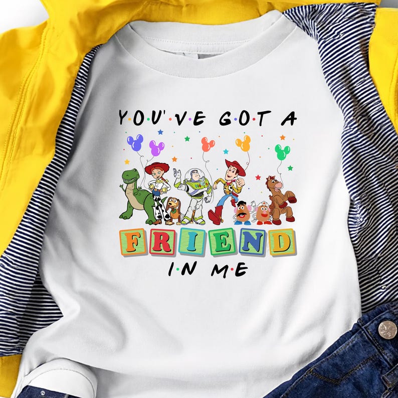 May include: White t-shirt featuring a colorful graphic of Toy Story characters with the text "YOU'VE GOT A FRIEND IN ME". The design includes Woody, Buzz Lightyear, and other characters from the movie, with balloons and stars.