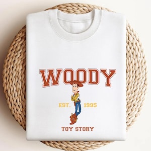 K&ouml;nnte beinhalten: Wei&szlig;es Sweatshirt mit einem Woody-Motiv aus Toy Story. Das Design zeigt das Wort "WOODY" in braunen Blockbuchstaben &uuml;ber einer Illustration der Figur, mit "EST. 1995" und "TOY STORY" darunter.