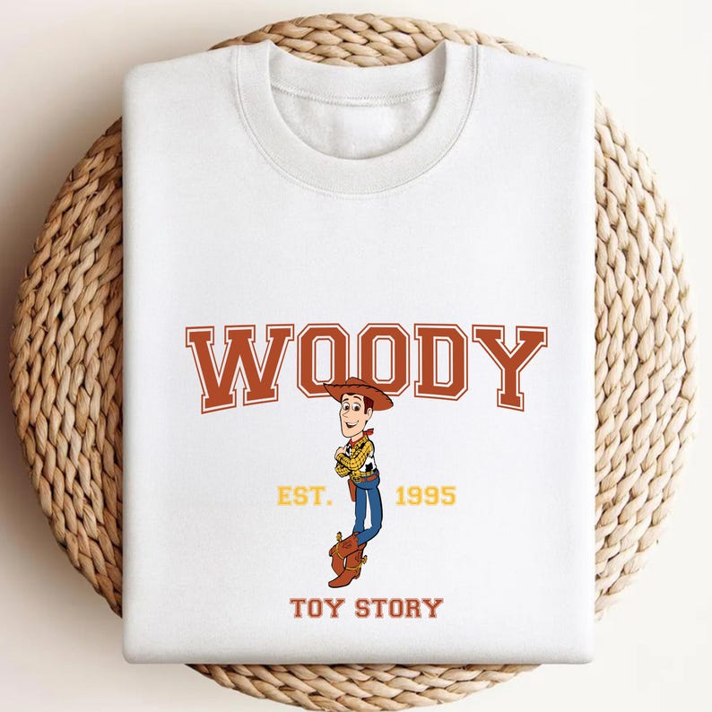 K&ouml;nnte beinhalten: Wei&szlig;es Sweatshirt mit einem Woody-Motiv aus Toy Story. Das Design zeigt das Wort "WOODY" in braunen Blockbuchstaben &uuml;ber einer Illustration der Figur, mit "EST. 1995" und "TOY STORY" darunter.