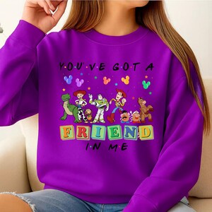 May include: A purple sweatshirt featuring the phrase "YOU'VE GOT A FRIEND IN ME" with cartoon character graphics. The design includes characters from a popular animated movie, with colorful block letters spelling out "FRIEND".
