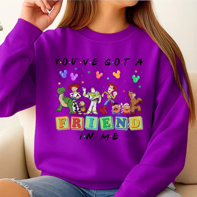 May include: A purple sweatshirt featuring the phrase "YOU'VE GOT A FRIEND IN ME" with cartoon character graphics. The design includes characters from a popular animated movie, with colorful block letters spelling out "FRIEND".