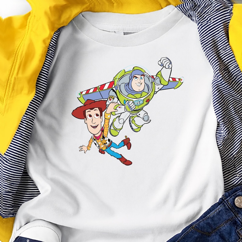 K&ouml;nnte beinhalten: Wei&szlig;es Raglan-&Auml;rmel-Shirt mit einer Cartoon-Grafik von Woody und Buzz Lightyear aus Toy Story. Woody tr&auml;gt einen Cowboyhut, und Buzz ist in einem Raumanzug mit Fl&uuml;geln. Das Shirt liegt auf einer gelben Jacke und einer blauen Jeans.