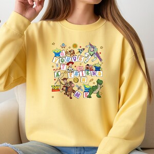 Puede incluir: Sudadera amarilla clara con un dise&ntilde;o gr&aacute;fico colorido de personajes de la pel&iacute;cula animada Toy Story. El dise&ntilde;o incluye la frase "You've Got a Friend in Me" y estrellas.