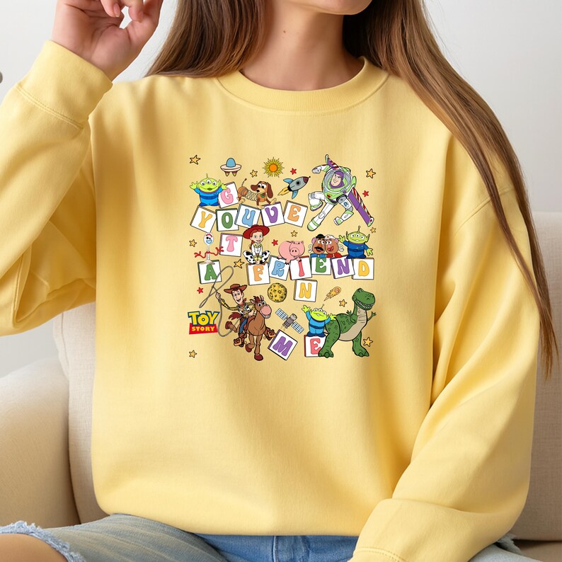 Puede incluir: Sudadera amarilla clara con un dise&ntilde;o gr&aacute;fico colorido de personajes de la pel&iacute;cula animada Toy Story. El dise&ntilde;o incluye la frase "You've Got a Friend in Me" y estrellas.