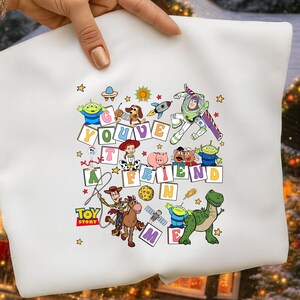 Puede incluir: Sudadera blanca con un gr&aacute;fico colorido de personajes de Toy Story y la frase "You've Got a Friend in Me". El dise&ntilde;o incluye a Woody, Buzz Lightyear y otros personajes de la pel&iacute;cula animada, rodeados de estrellas.