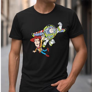 K&ouml;nnte beinhalten: Schwarzes T-Shirt mit einer farbenfrohen Grafik von Woody und Buzz Lightyear aus Toy Story. Woody tr&auml;gt einen Cowboyhut und Buzz einen Raumanzug. Das Design ist im lebendigen Cartoon-Stil gehalten.