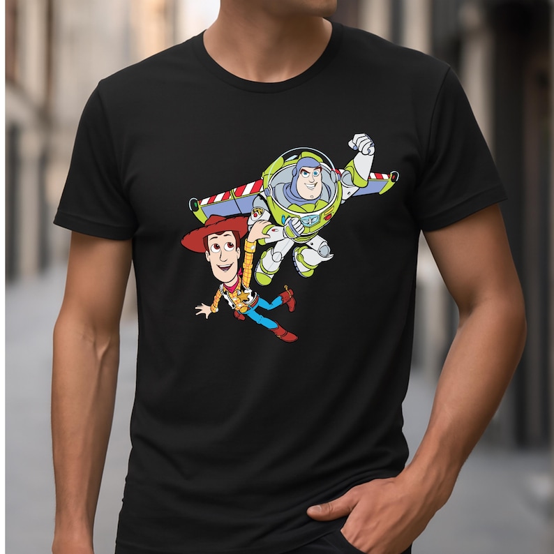 K&ouml;nnte beinhalten: Schwarzes T-Shirt mit einer farbenfrohen Grafik von Woody und Buzz Lightyear aus Toy Story. Woody tr&auml;gt einen Cowboyhut und Buzz einen Raumanzug. Das Design ist im lebendigen Cartoon-Stil gehalten.