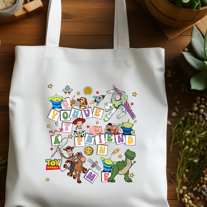 Puede incluir: Bolso tote blanco con un gr&aacute;fico colorido de personajes de la pel&iacute;cula animada "Toy Story". El dise&ntilde;o incluye la frase "You've Got a Friend in Me" con varios personajes y estrellas.