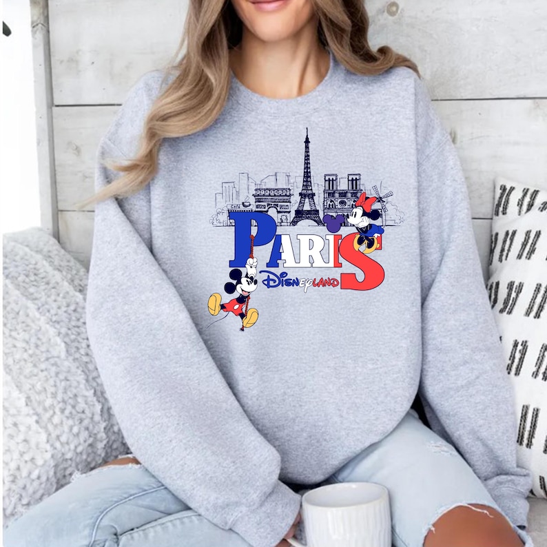 May include: A light grey sweatshirt with a graphic of the Paris skyline, the word "PARIS" in blue and red, and two Mickey Mouse characters. The text "Disneyland" is below the word "PARIS".